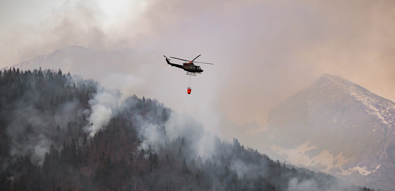 Firefighting helicopter with a bucket suspended on a cable, collecting water from a mountain lake to extinguish the flame in a forest