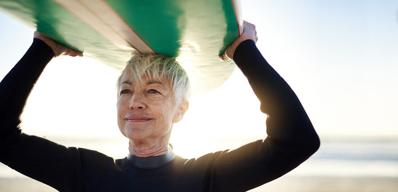 mature female surfer carrying surfboard over head.
