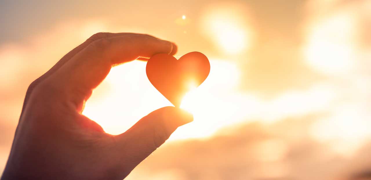 A hand holding a heart-shaped object up to the sun.