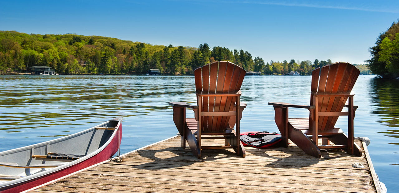 Two Muskoka chairs on a wooden dock overlooking the blue water of a lake in Muskoka, Ontario Canada. A red canoe is tied to the pier and life jackets are visible near the chairs.